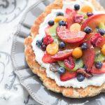 Pizza-déjeuner avec une pâte à pizza dorée, garnie de fromage frais et d'une variété de fruits frais. Le tout déposé sur une assiette grise sur un fond blanc et gris.
