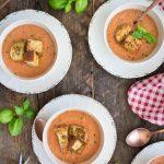 Un potage froid et frais à base de tomates, concombre et de pain baguette. La soupe est de couleur orangé, décoré de croûtons de pain dans un bol blanc. Le tout déposé sur un fond de couleur bois.