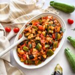 Ratatouille à base d'aubergine, de courgettes, poivrons et de fenouil. Le tout déposé sur une assiette blanche ovale, une cuillère à mélanger blanche. L'assiette est déposé à côté d'un linge de vaisselle beige et blanc.