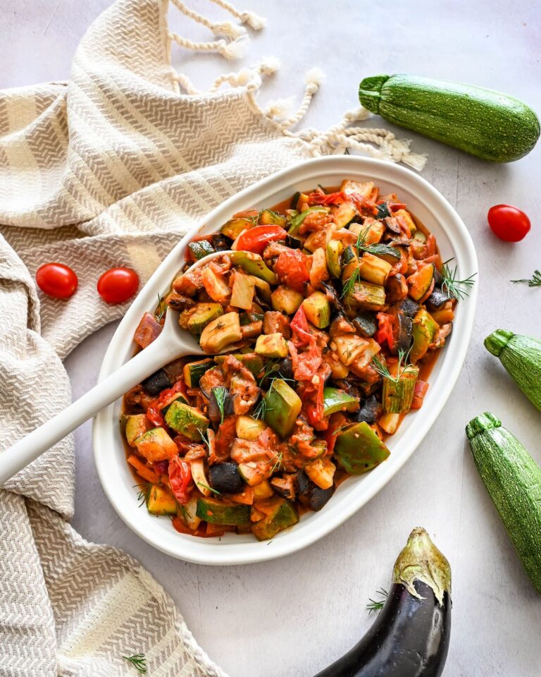 Ratatouille à base d'aubergine, de courgettes, poivrons et de fenouil. Le tout déposé sur une assiette blanche ovale, une cuillère à mélanger blanche. L'assiette est déposé à côté d'un linge de vaisselle beige et blanc.