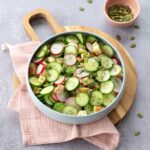 Salade de concombre, radis et pomme dans un bol lilas déposé sur une planche de bois ronde et un linge de vaisselle vieux rose.