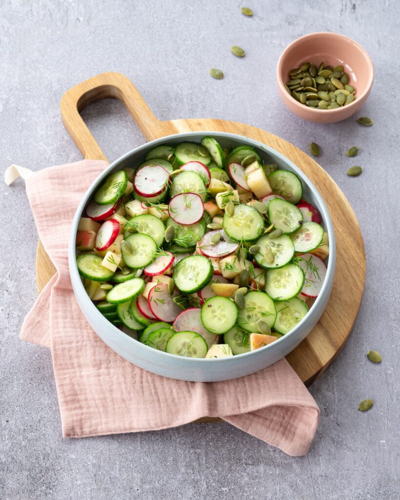 Salade de concombre, radis et pomme dans un bol lilas déposé sur une planche de bois ronde et un linge de vaisselle vieux rose.