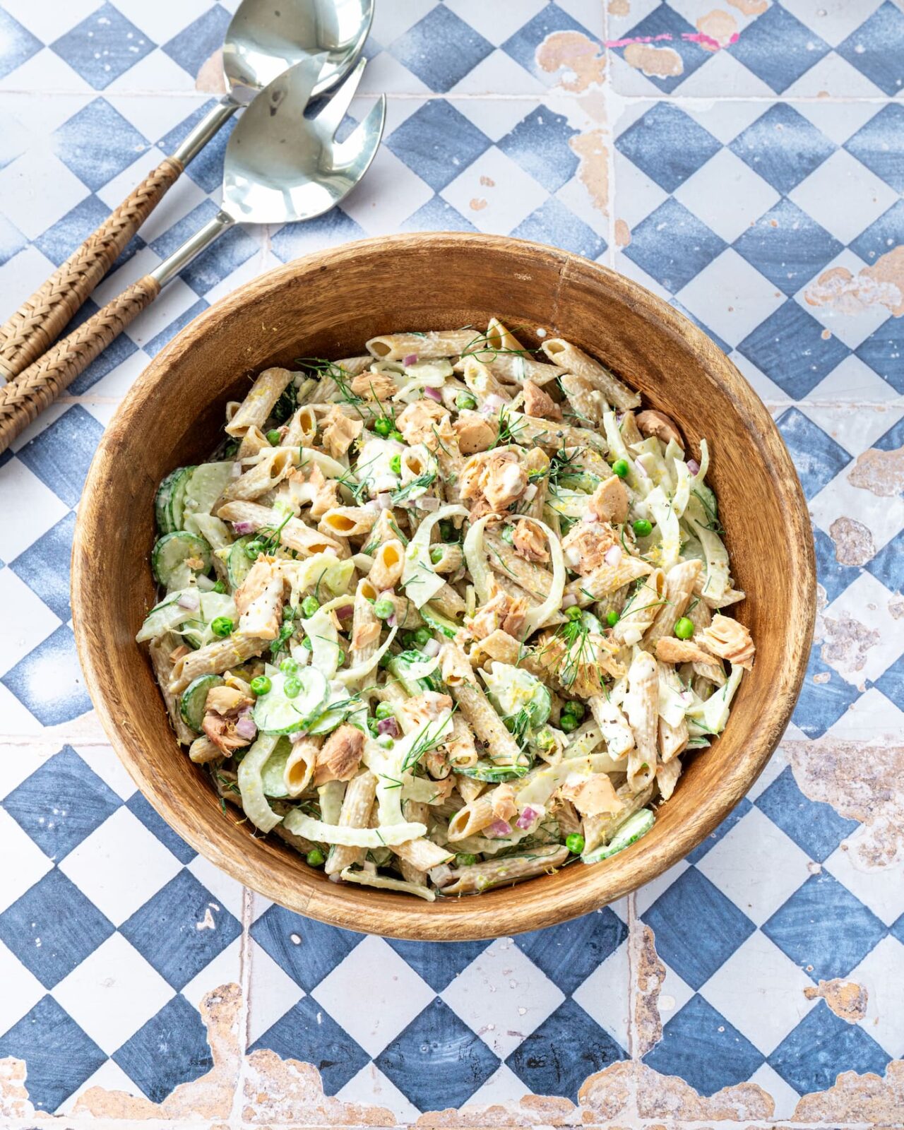 Salade de pâtes et saumon, fenouil, concombre et petits pois dans un bol en bois déposé sur un fond en losange bleu et blanc.