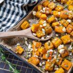 Jolis cubes de citrouille rôtis déposés sur une plaque de cuisson noir et parsemés de feta et de graines de citrouille