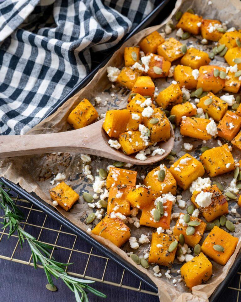 Jolis cubes de citrouille rôtis déposés sur une plaque de cuisson noir et parsemés de feta et de graines de citrouille