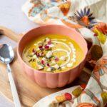 Magnifique potage à la citrouille déposé dans un bol de couleur rose-orangé, garnit de morceaux de pomme et de graines de citrouille