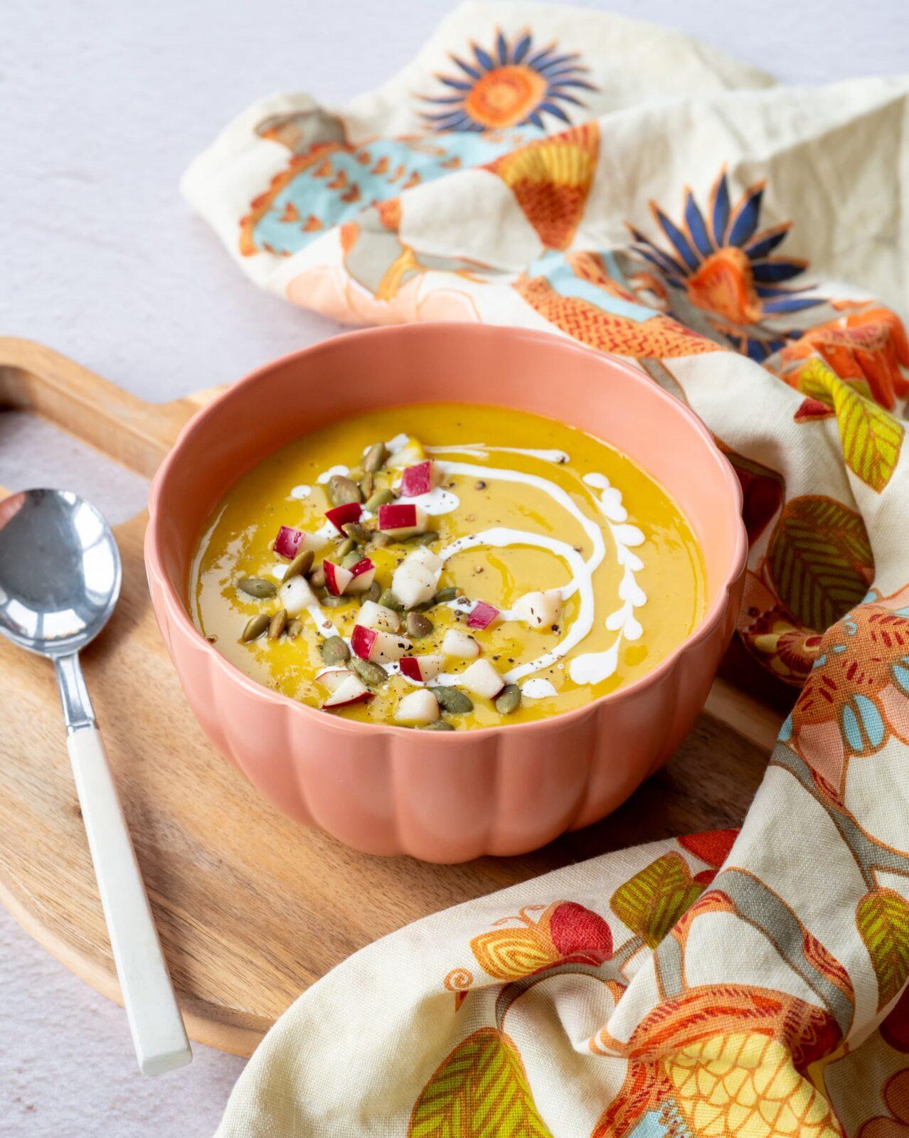 Magnifique potage à la citrouille déposé dans un bol de couleur rose-orangé, garnit de morceaux de pomme et de graines de citrouille