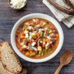 Bol de soupe rempli de légumes, accompagné de deux tranches de pain, de parmesan et de deux cuillères