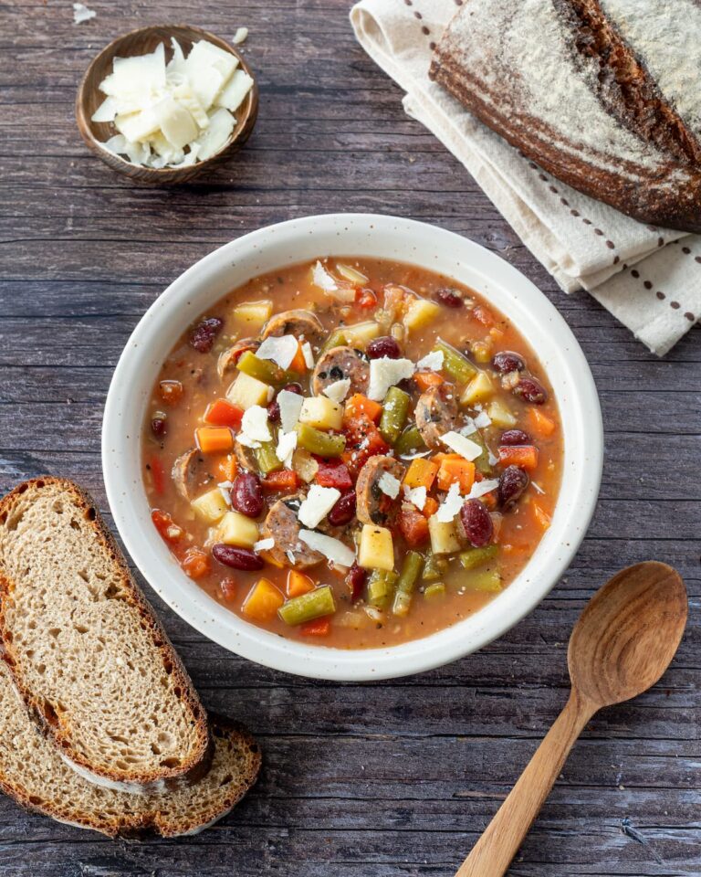 Bol de soupe rempli de légumes, accompagné de deux tranches de pain, de parmesan et de deux cuillères