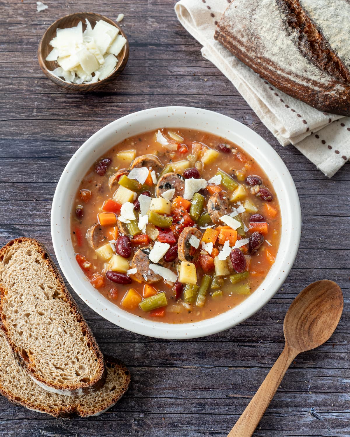 Bol de soupe rempli de légumes, accompagné de deux tranches de pain, de parmesan et de deux cuillères