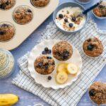 Une petite assiette avec 2 muffins aux bananes et bleuets, accompagnée de tranches de bananes et d'un bol de yogourt