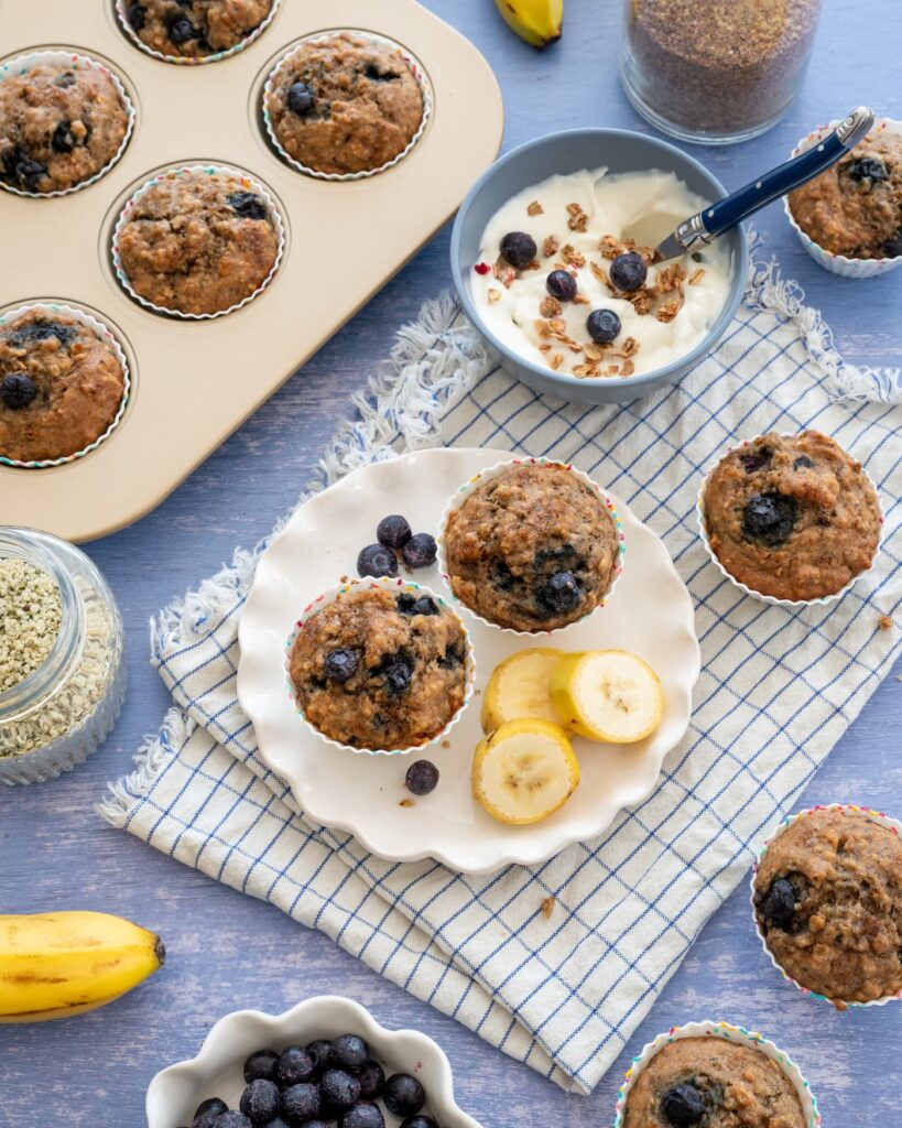 Une petite assiette avec 2 muffins aux bananes et bleuets, accompagnée de tranches de bananes et d'un bol de yogourt