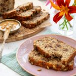2 tranches de pain aux bananes et chocolat déposées dans une assiette rose et accompagnées de jolies fleurs dans un pot
