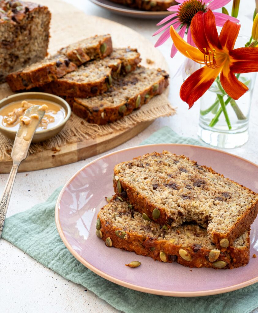 2 tranches de pain aux bananes et chocolat déposées dans une assiette rose et accompagnées de jolies fleurs dans un pot