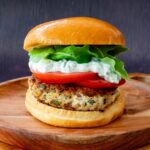 Burger de porc aux herbes et feta garnit de sauce, de roquette et de tomate, déposé sur une assiette en bois