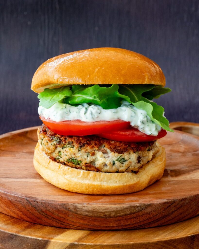 Burger de porc aux herbes et feta garnit de sauce, de roquette et de tomate, déposé sur une assiette en bois