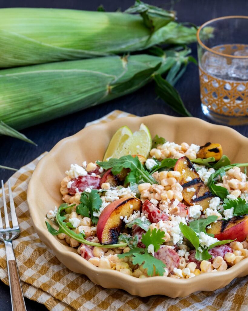 Salade de maïs cru aux saveurs Tex-Mex servie dans un bol jaune et accompagnée de maïs entier et d'une verre d'eau